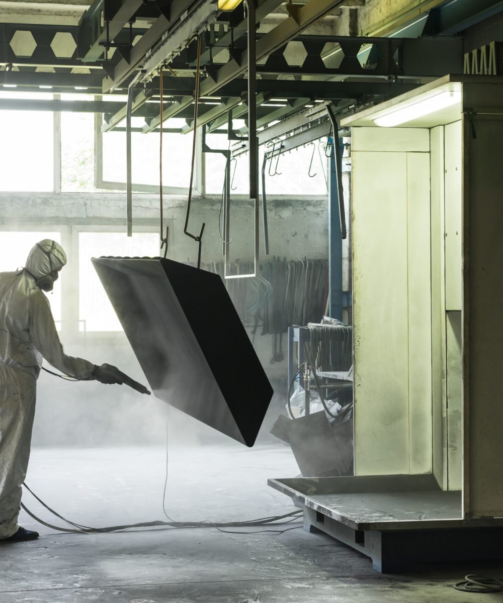 view of a worker wearing a full white protective suit and breathing mask, sand blasting a metal crate hung from a metal beam in the ceiling of an industrial hall