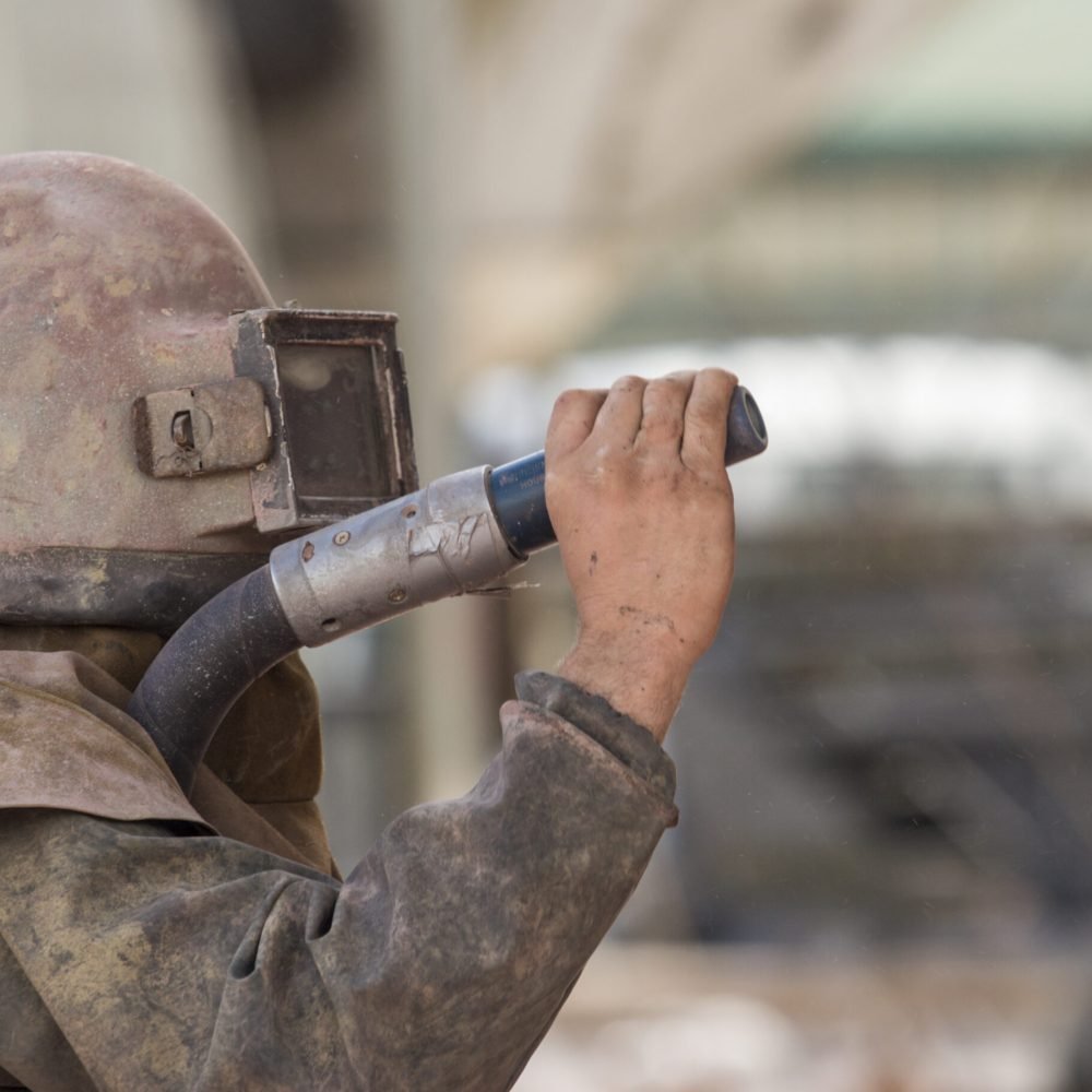 worker, sandblasting the corroded hull of a sailing vessel with a high pressure sandblasting syst