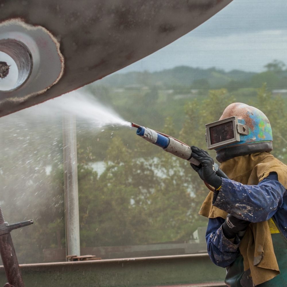 worker, sandblasting the corroded hull of a sailing vessel with a high pressure sandblasting syst